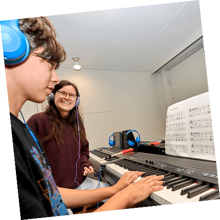 A piano teacher smiles at a teen student as he plays on a keyboard during a piano lesson.