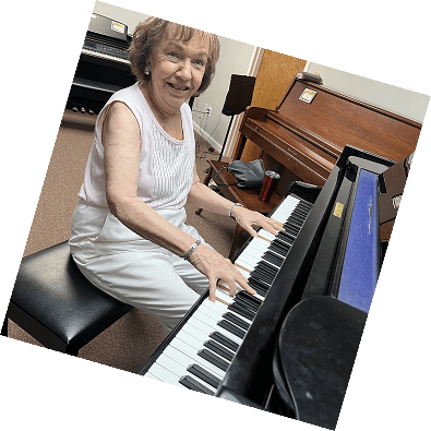 An elderly woman smiles as she practices a popular song on a wooden acoustic grand piano.